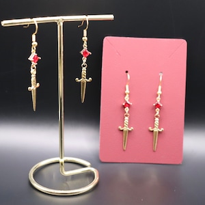 May include: Four gold dagger earrings with red gemstone accents. The earrings are displayed on a gold stand and a pink card.