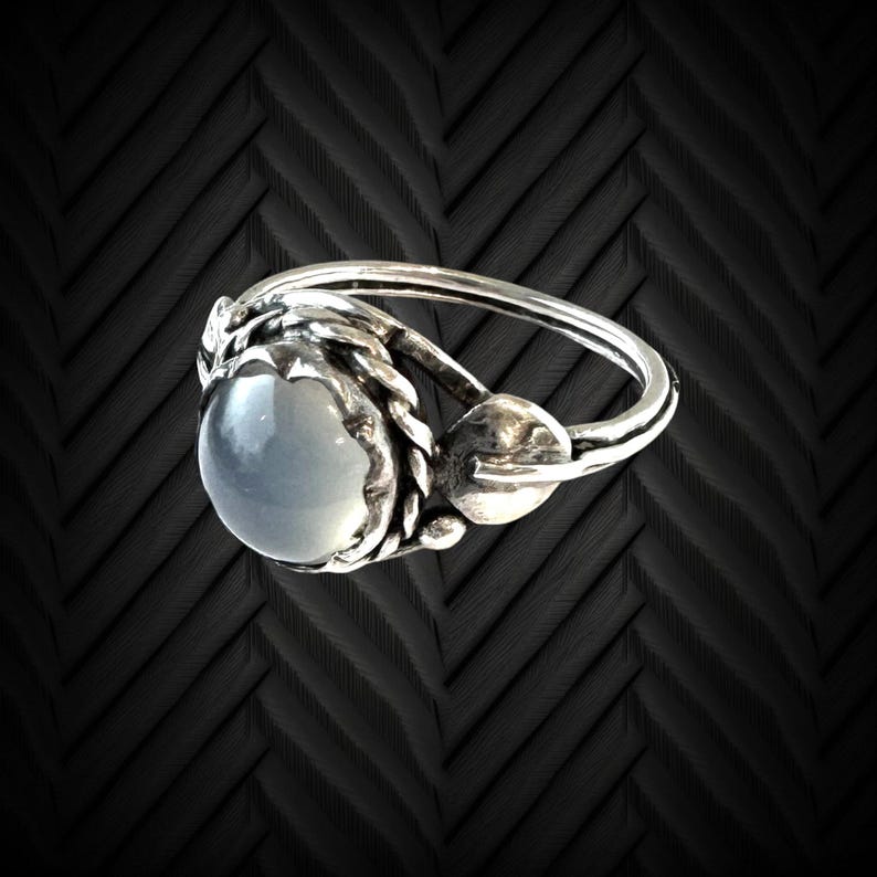 May include: A silver ring with a large, round, pale blue gemstone. The ring has a detailed, textured design around the gemstone and on the band. The ring is set against a dark background.