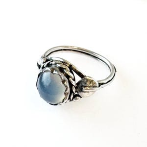 May include: A silver ring featuring a light blue, oval-shaped gemstone. The ring has a detailed, textured design around the stone and on the band, with a floral or leaf-like accent on the side. The ring is set against a white background.