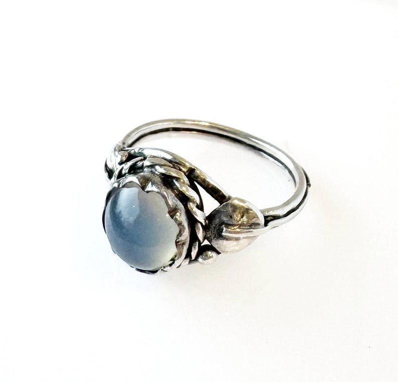May include: A silver ring featuring a light blue, oval-shaped gemstone. The ring has a detailed, textured design around the stone and on the band, with a floral or leaf-like accent on the side. The ring is set against a white background.