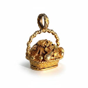 May include: A gold-colored pendant shaped like a basket, intricately detailed with grapes, leaves, and fruit. The basket has a twisted handle and is likely a charm or small jewelry piece. The pendant is on a white background.
