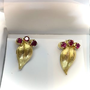 May include: A pair of gold-coloured earrings with a leaf design. Each earring features three red gemstones at the top and a textured leaf shape. The earrings are displayed on a white surface.