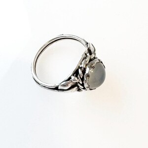 May include: A silver ring with a light gray, oval-shaped stone. The ring features a detailed leaf design around the stone and along the band. The ring is set against a white background, highlighting its intricate details.