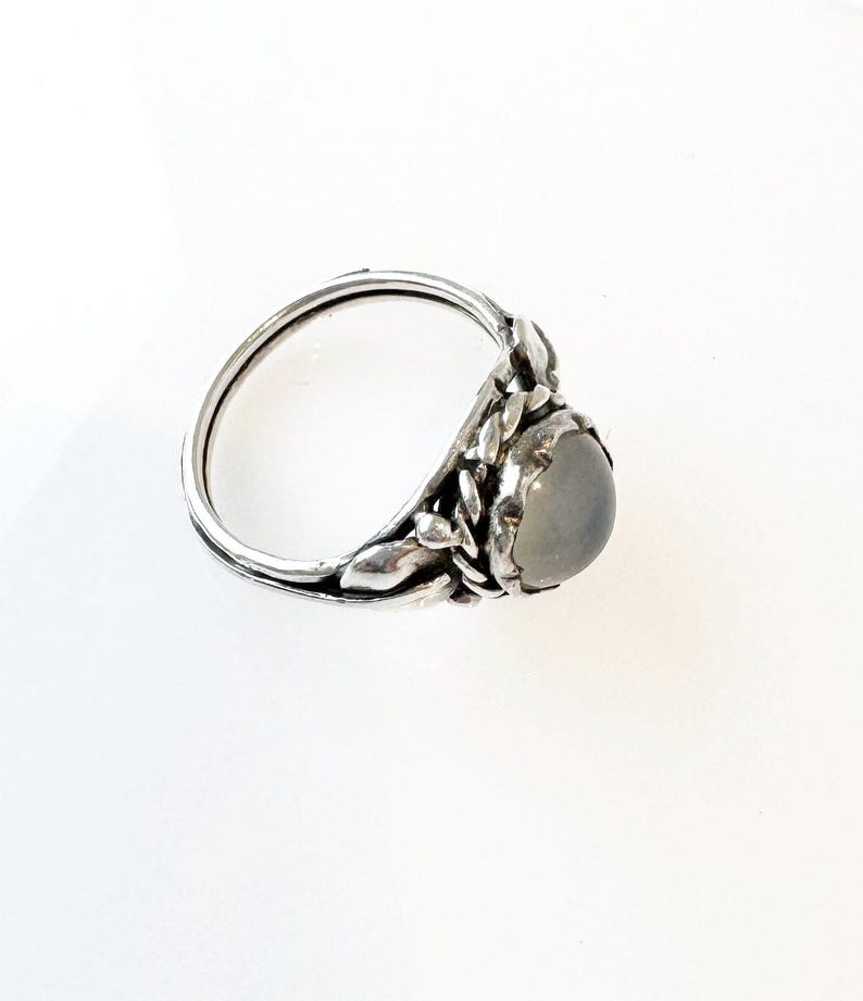 May include: A silver ring with a light gray, oval-shaped stone. The ring features a detailed leaf design around the stone and along the band. The ring is set against a white background, highlighting its intricate details.