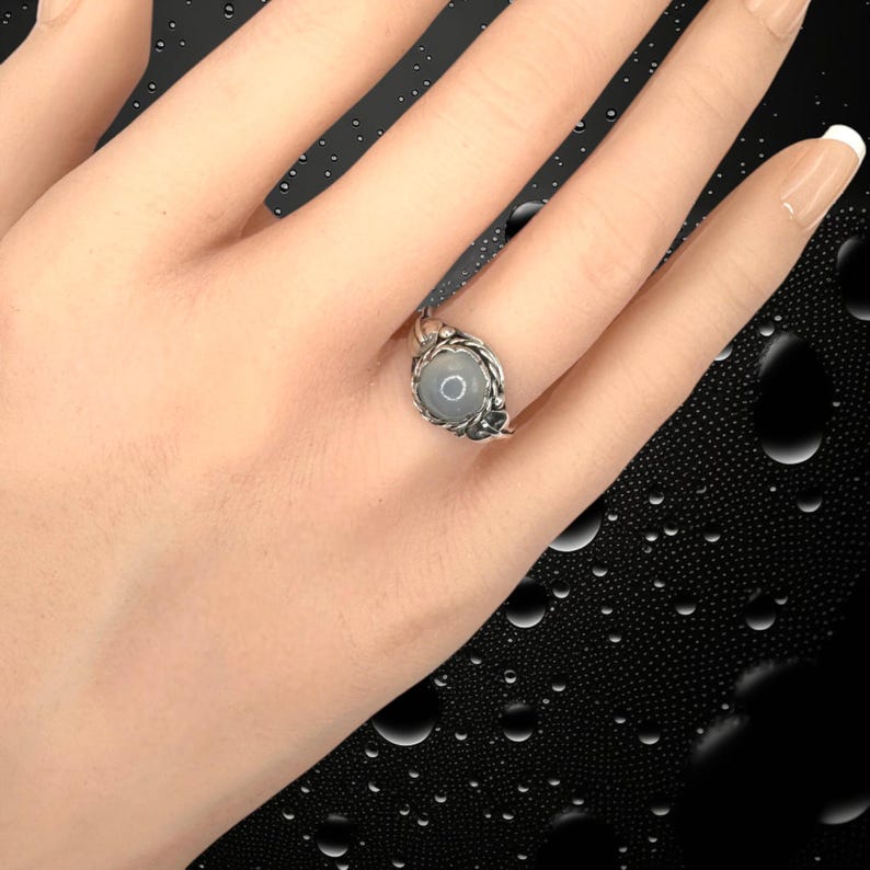 May include: A silver ring with a round, light gray stone set in a decorative, twisted metal band. The ring is on a person's finger, with a black background.