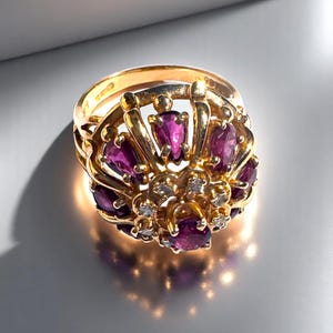 May include: A gold ring featuring a cluster of pear-shaped deep pink gemstones arranged in a radial pattern, accented with small clear stones in the centre. The metal band has a polished finish and visible hallmarks on the inner surface.