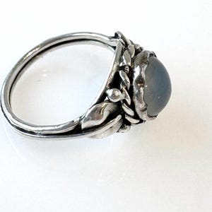 May include: A silver ring with a large, oval, gray gemstone. The ring features a floral design around the gemstone, with detailed leaves and small buds. The band is smooth and polished, creating a contrast with the textured setting.