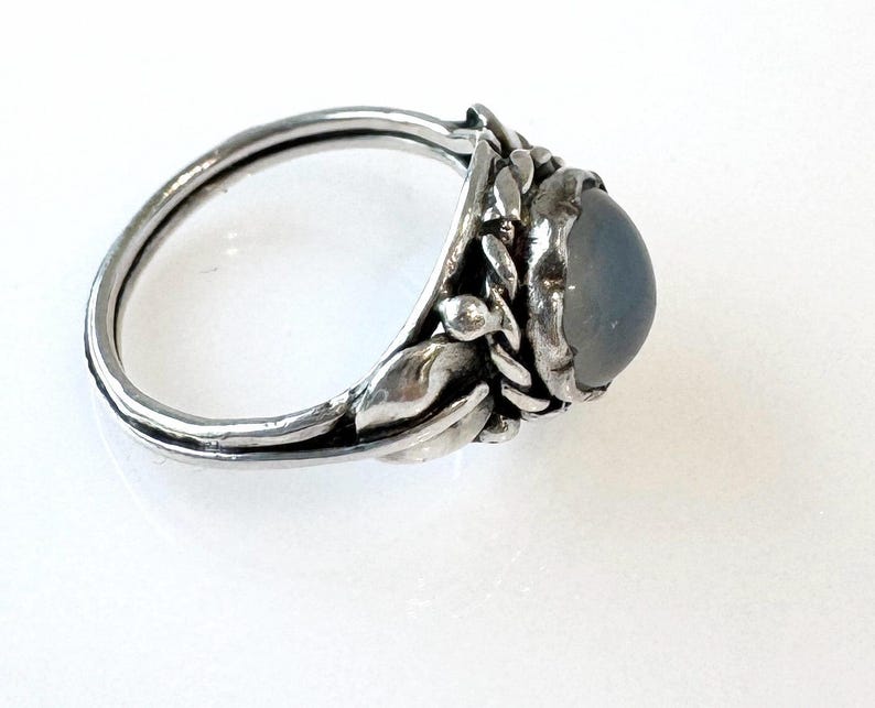 May include: A silver ring with a large, oval, gray gemstone. The ring features a floral design around the gemstone, with detailed leaves and small buds. The band is smooth and polished, creating a contrast with the textured setting.