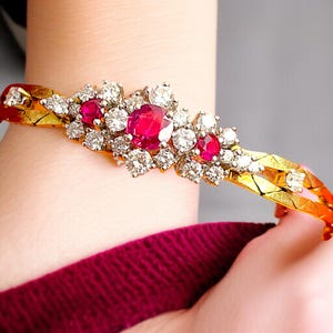 May include: A gold bracelet with a central cluster of diamonds and red rubies. The bracelet's band has a textured design. The jewellery is worn on a wrist, highlighting the contrast of the gemstones and the gold's shine.