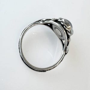 May include: A silver ring with an oval, dark-colored stone. The ring features a detailed, ornate design around the stone and along the band. The band has a delicate, floral-like pattern. The ring is set against a white background.