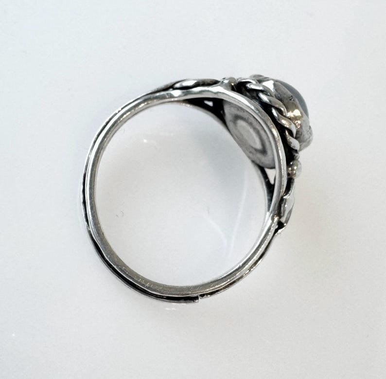 May include: A silver ring with an oval, dark-colored stone. The ring features a detailed, ornate design around the stone and along the band. The band has a delicate, floral-like pattern. The ring is set against a white background.