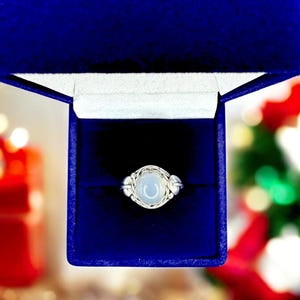 May include: A silver ring with a light blue, oval-shaped stone, presented in a royal blue jewelry box. The ring features a woven design around the stone and small decorative elements on either side. The box is open, revealing the ring against a dark interior.