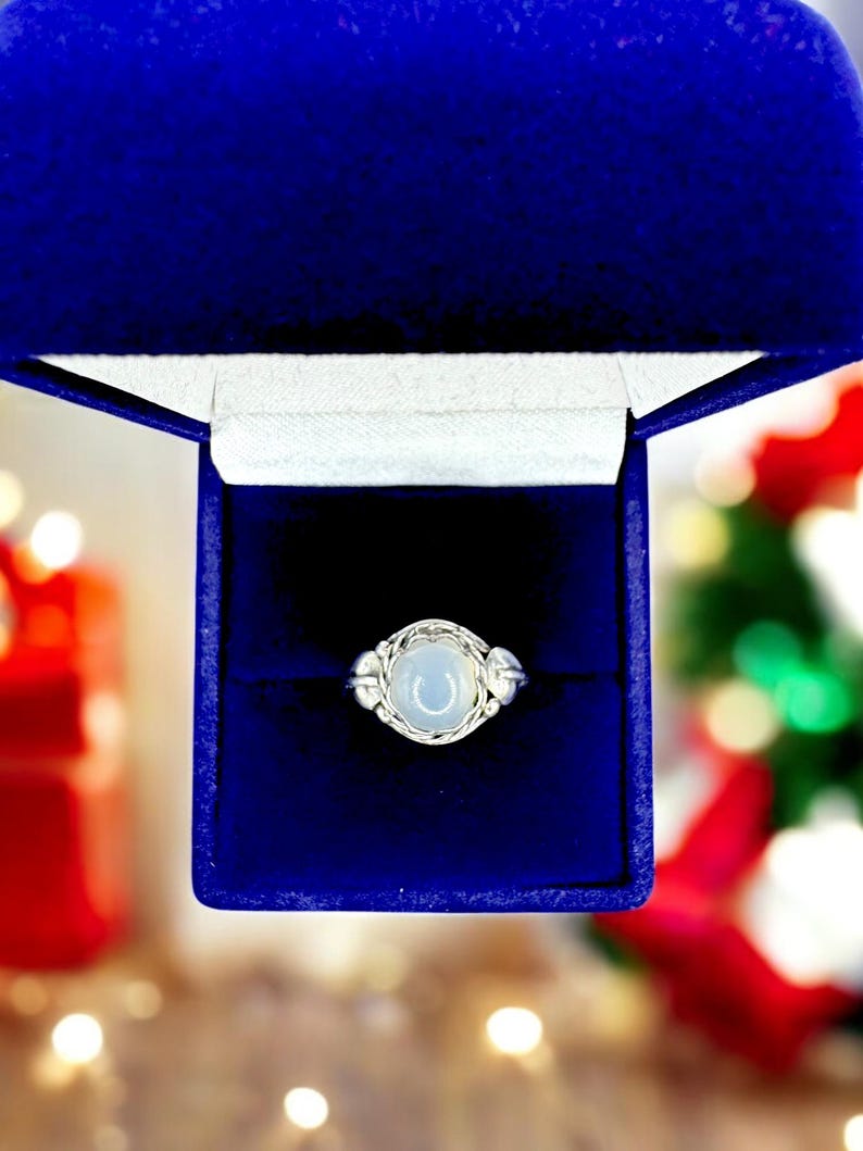 May include: A silver ring with a light blue, oval-shaped stone, presented in a royal blue jewelry box. The ring features a woven design around the stone and small decorative elements on either side. The box is open, revealing the ring against a dark interior.