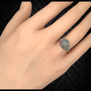 May include: A silver ring with a round, light blue gemstone set in a decorative, twisted metal band. The ring is on a person's finger, showcasing the intricate design and the stone's subtle color. The ring is a piece of jewelry.