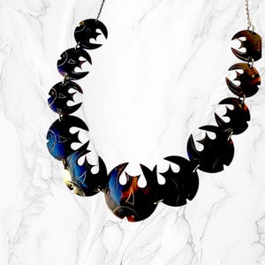 May include: A dark metal necklace featuring a repeating fish design. Each fish has a crescent shape with cut-out details and a gradient of blue, red, and gold hues. The necklace hangs on a delicate chain against a white marble background.