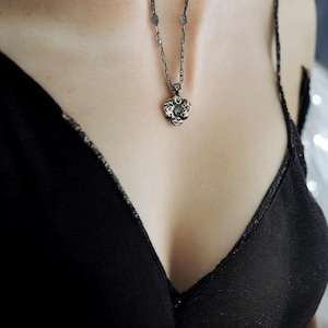 May include: A silver-toned necklace with a floral pendant. The pendant is detailed with intricate patterns and a central stone. The necklace is worn against a black top, creating a contrast. The chain is delicate and complements the pendant's design.