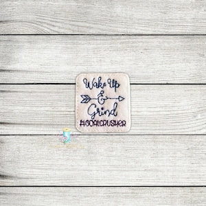 Puede incluir: Parche de fieltro beige con bordado negro que dice "Wake Up & Grind #GoalCrusher". Una flecha está bordada sobre el texto.