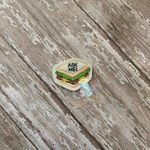 May include: A white felt patch with an embroidered image of a sandwich with the text "ASK ME" on top.