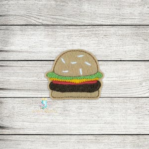 May include: A felt burger applique with a tan bun, green lettuce, yellow cheese, red tomato, and a dark brown patty. The burger has white sprinkles and is stitched with a brown thread. The applique is on a white wooden background.