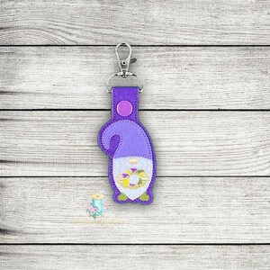 May include: Purple felt gnome keyring with a wreath of flowers in its hands. The gnome is wearing a purple hat and has a white beard. The keyring has a silver metal ring.