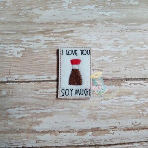Può includere: Carta di feltro bianco con il testo "Ti amo molto soia" e un'immagine di cartone animato di una bottiglia di salsa di soia.