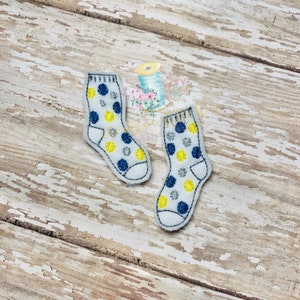 Puede incluir: Dos calcetines de fieltro blanco con lunares azules, amarillos y plateados. Los calcetines están bordados y tienen un fondo blanco.
