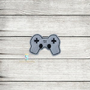 May include: A gray felt video game controller with black stitching and black plus and circle buttons. The controller is centered on a light wood grain background.