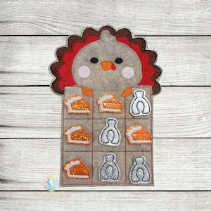 May include: A felt turkey game with a brown turkey head and red feathers. The game board has nine squares with images of pie slices and turkey feet.