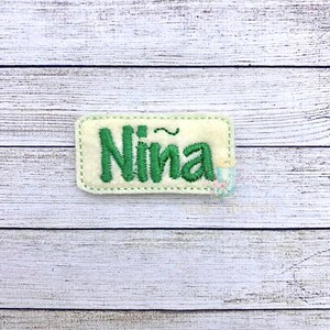 May include: A white felt name tag with green embroidered lettering that reads "Nina" with a tilde over the "n".