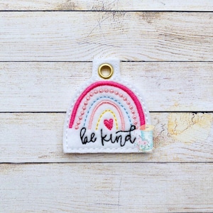 May include: A white felt keychain with a rainbow design and the words "be kind" embroidered in black. A small pink heart is in the centre of the rainbow.