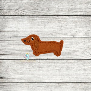 Peut inclure: Une applique en feutre marron en forme de dachshund avec un visage blanc et des yeux noirs.