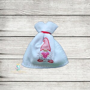May include: A white fabric bag with a red drawstring and an embroidered gnome with a pink heart. The gnome is wearing a red hat with white stripes.
