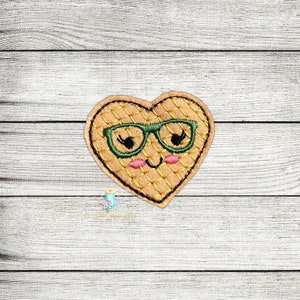 Könnte beinhalten: Eine herzförmige Waffel mit einem lächelnden Gesicht, die eine grüne Brille trägt. Die Waffel ist gelb mit einem braunen Gittermuster.