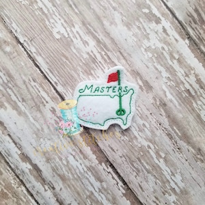 Op de afbeelding: Groene vilt omtrek van de Verenigde Staten met een rode vlag en een golfclub, met het woord "MASTERS" in witte letters.