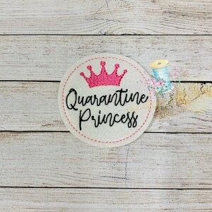May include: A white circle with a pink crown and the text "Quarantine Princess" in black cursive lettering.