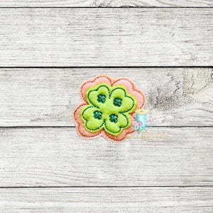 Può includere: Un quadrifoglio verde con contorno rosa. Il quadrifoglio è ricamato su una superficie di legno bianco.