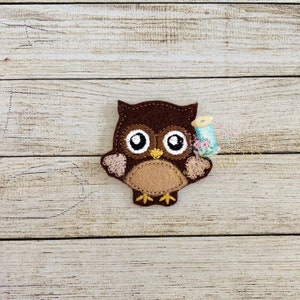 May include: A brown felt owl with large, white eyes and a yellow beak. The owl is standing on a wooden surface.