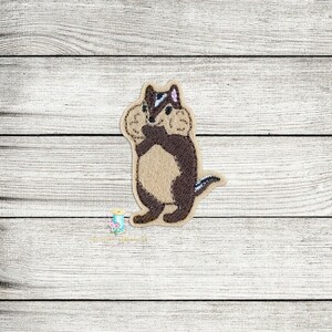 May include: Embroidered brown and white chipmunk with a pink nose, holding a nut in its paws. The chipmunk is standing on a white wooden background.