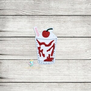 May include: Embroidered patch of a red and white milkshake with a cherry on top and a pink straw.