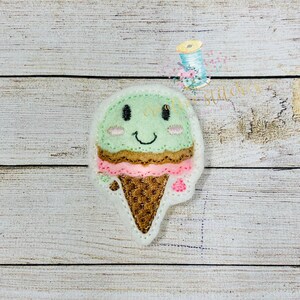 Pode incluir: Um cone de sorvete verde com um rosto sorridente, cobertura de chocolate marrom, granulado rosa e um cone de waffle.