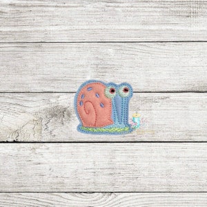 Peut inclure: Un escargot brodé rose et bleu avec de grands yeux. L'escargot est assis sur une base verte.