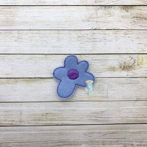 Könnte beinhalten: Eine hellblaue Filzblume mit einem violetten Zentrum. Die Blume befindet sich auf einem hellbraunen Holzhintergrund.