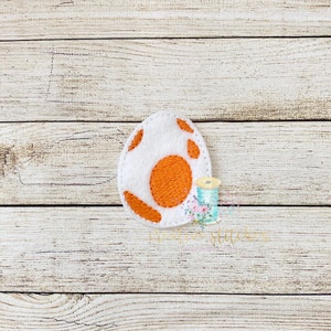 Peut inclure: Un œuf en feutre blanc avec un jaune d'œuf orange et des taches brunes. L'œuf est sur un fond en bois.