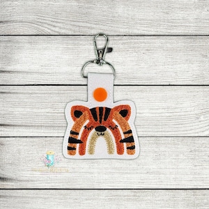 Pode incluir: Chaveiro de tecido branco com um lindo design de rosto de tigre laranja e preto. O tigre tem os olhos fechados e um nariz arco-íris. O chaveiro tem um anel de metal e um pequeno botão laranja.