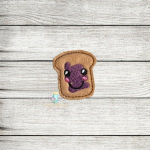 Peut inclure: Un patch brodé en feutre marron représentant une tranche de pain grillé avec de la confiture violette et un visage souriant.