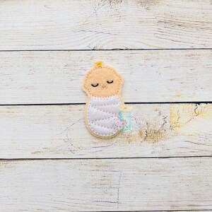 Puede incluir: Una aplicaci&oacute;n de fieltro de un beb&eacute; envuelto en una manta rosa con una cinta amarilla. El beb&eacute; tiene los ojos cerrados y una sonrisa cosida.