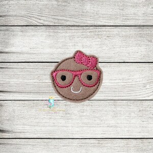 May include: A brown felt applique of a smiling face with pink glasses and a pink bow.