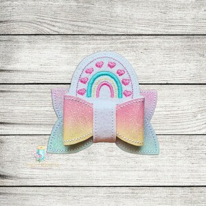 May include: A white, pink and yellow glitter bow with a white centre and a rainbow with pink hearts embroidered on top.