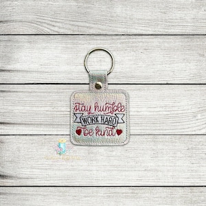 May include: A silver iridescent keychain with a square tag that reads "stay humble work hard be kind" in red embroidery. Two small red hearts are embroidered on either side of the text.