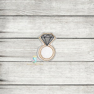 May include: A white felt ring with a grey diamond on top. The ring is outlined in gold thread.
